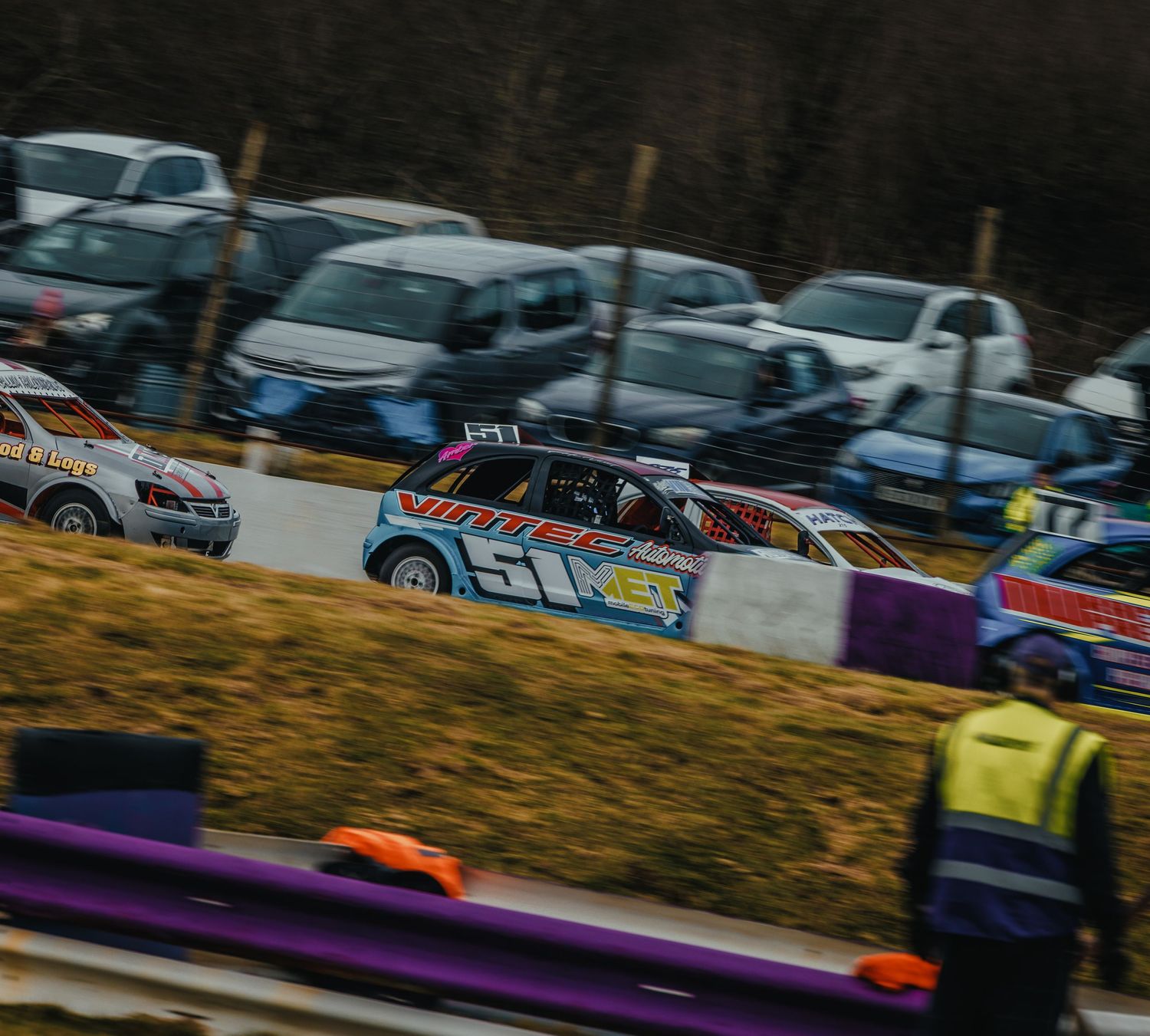 Multiple stock cars race around a dirt track corner with colorful barriers visible.