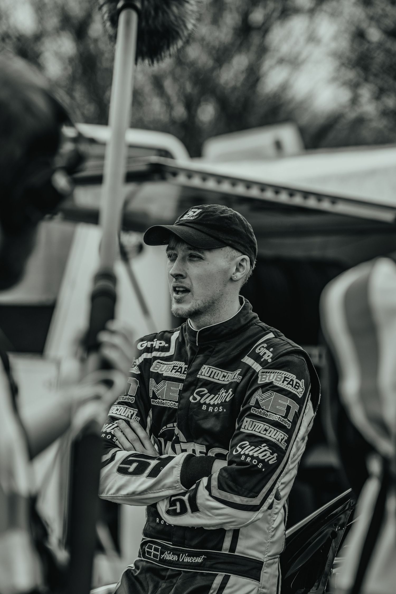 Racing driver in sponsor-covered uniform speaking at motorsport event.