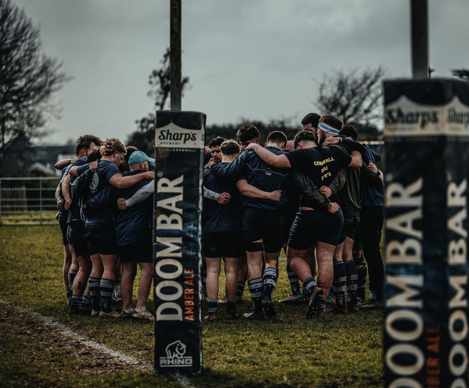 Rugby team huddles together before a match near field goal posts during an overcast day.