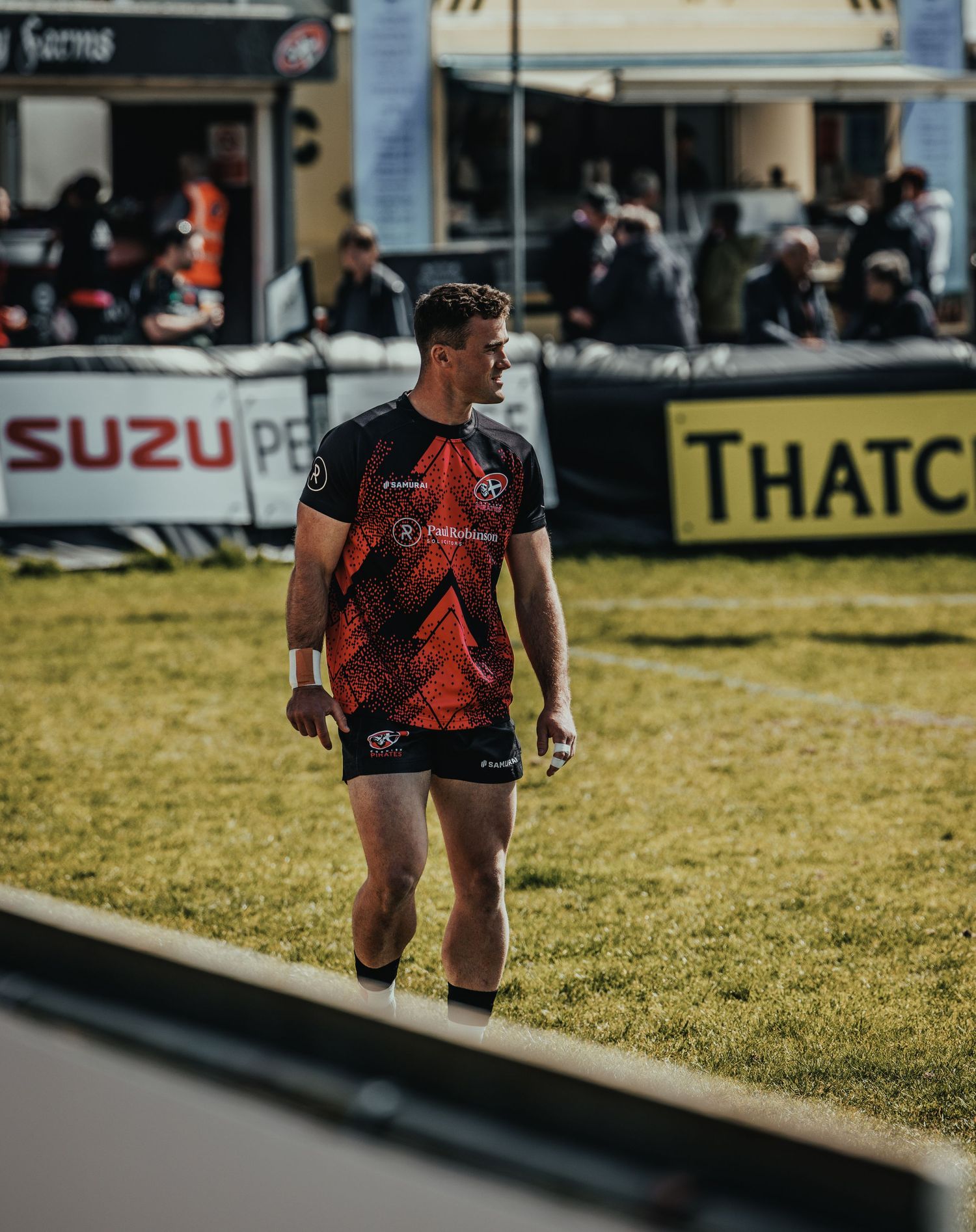 Rugby player in red and black jersey stands on field with Suzuki advertising banner visible.