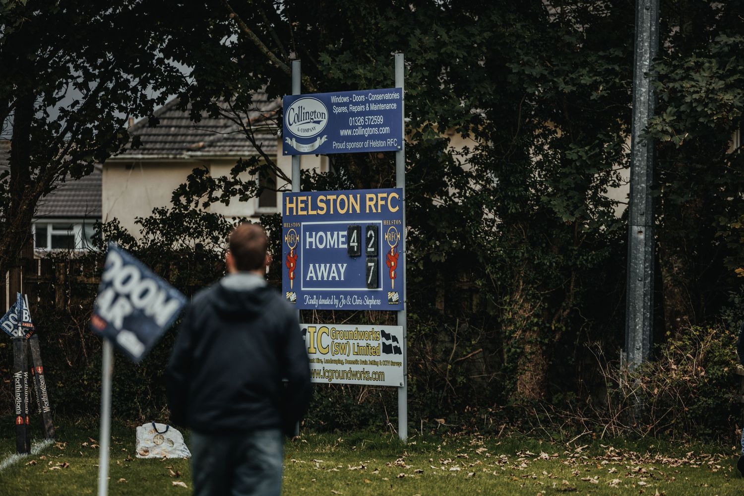 A person walks past the Helston RFC rugby club scoreboard sign showing a home match result outdoors.
