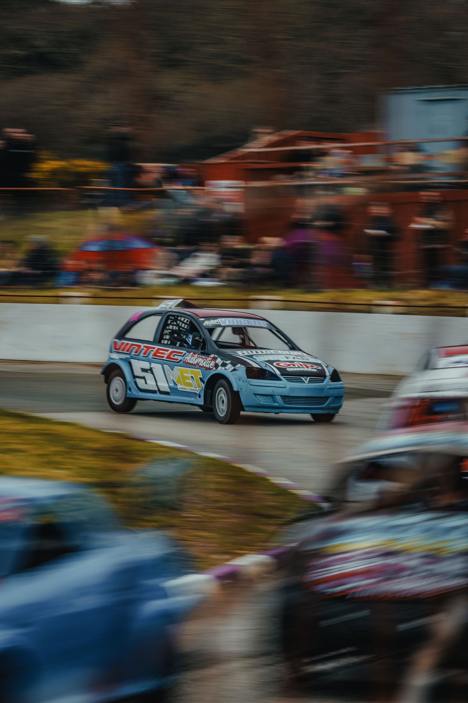 Blue race car competing on track with motion blur and spectators in background.