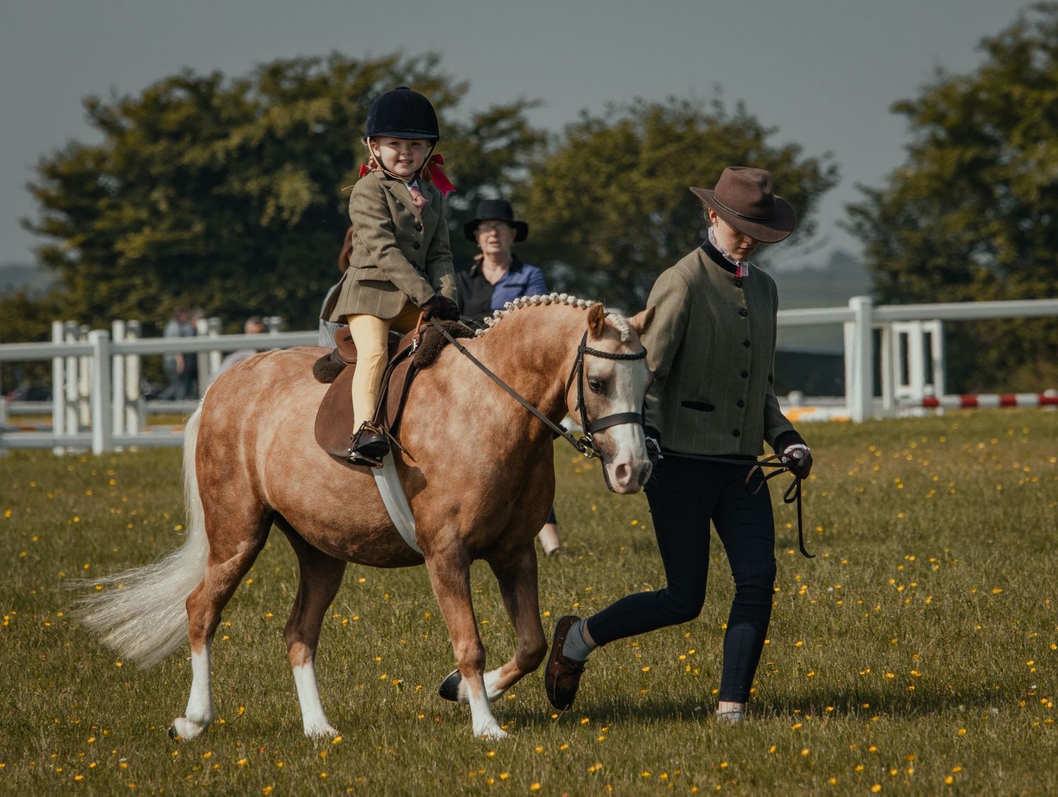 Child riding a palomino pony led by handler in an equestrian arena.
