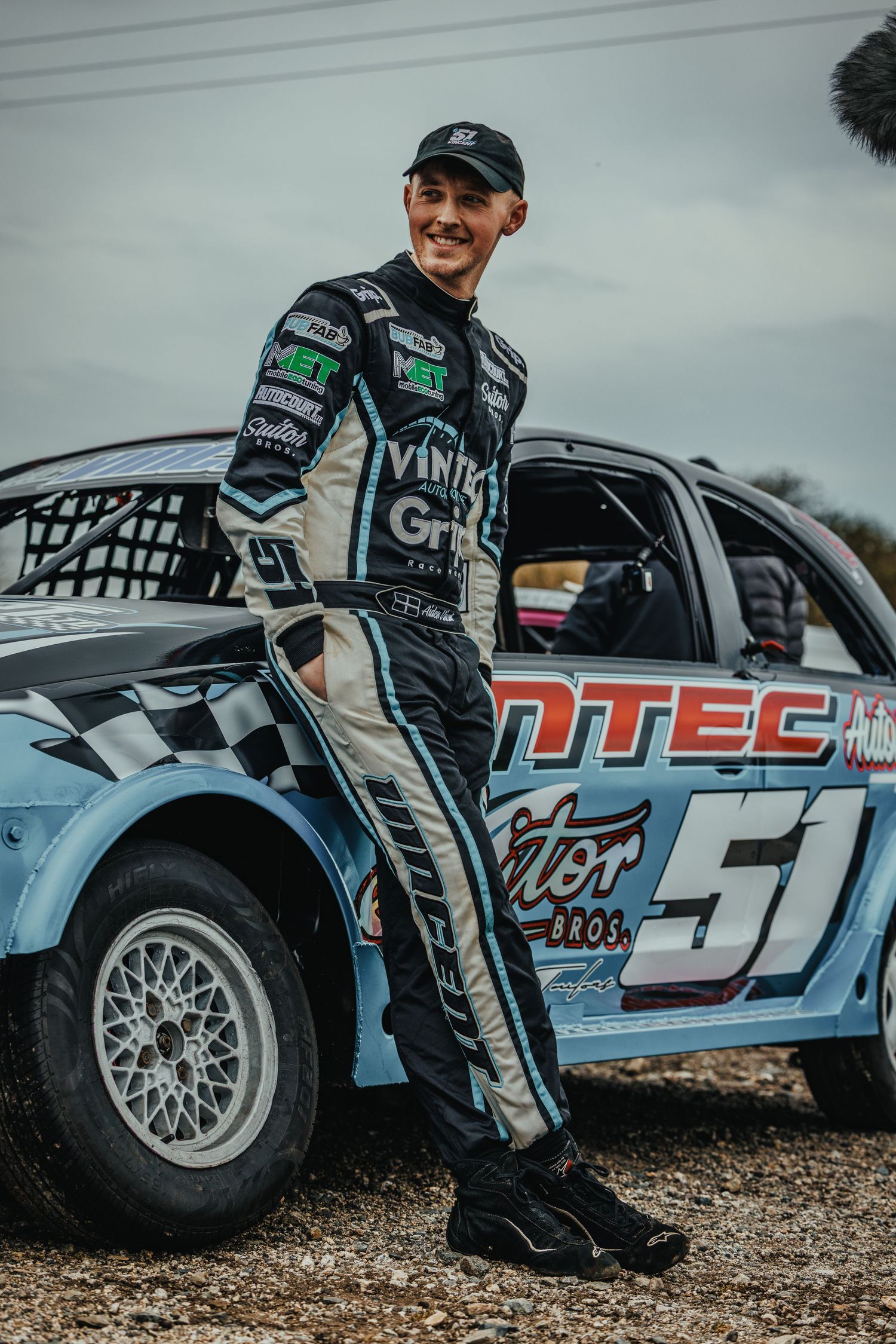 Racing driver in Vintec uniform poses confidently beside number 51 race car.