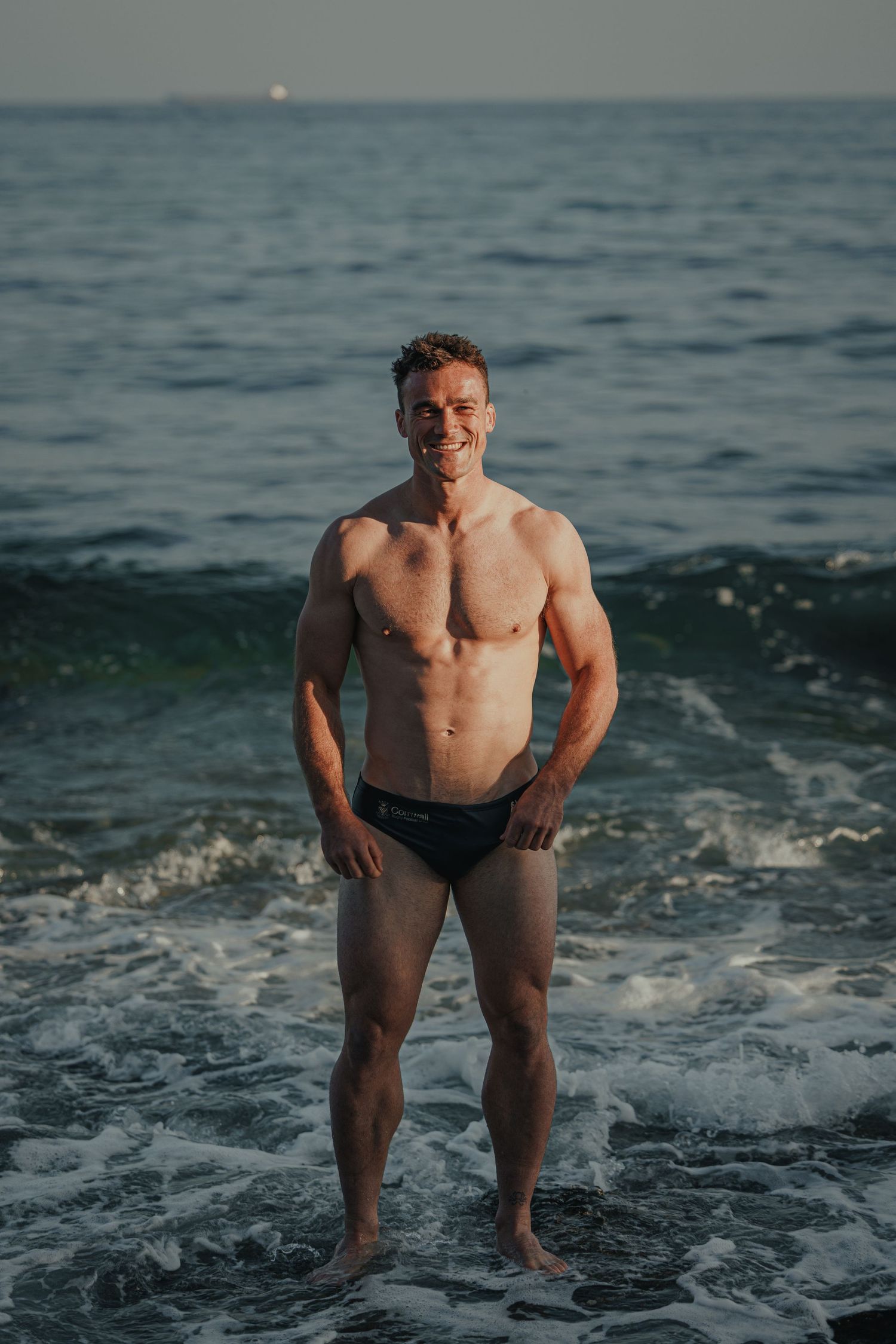 Fitness model posing in swim shorts against ocean backdrop at sunset.