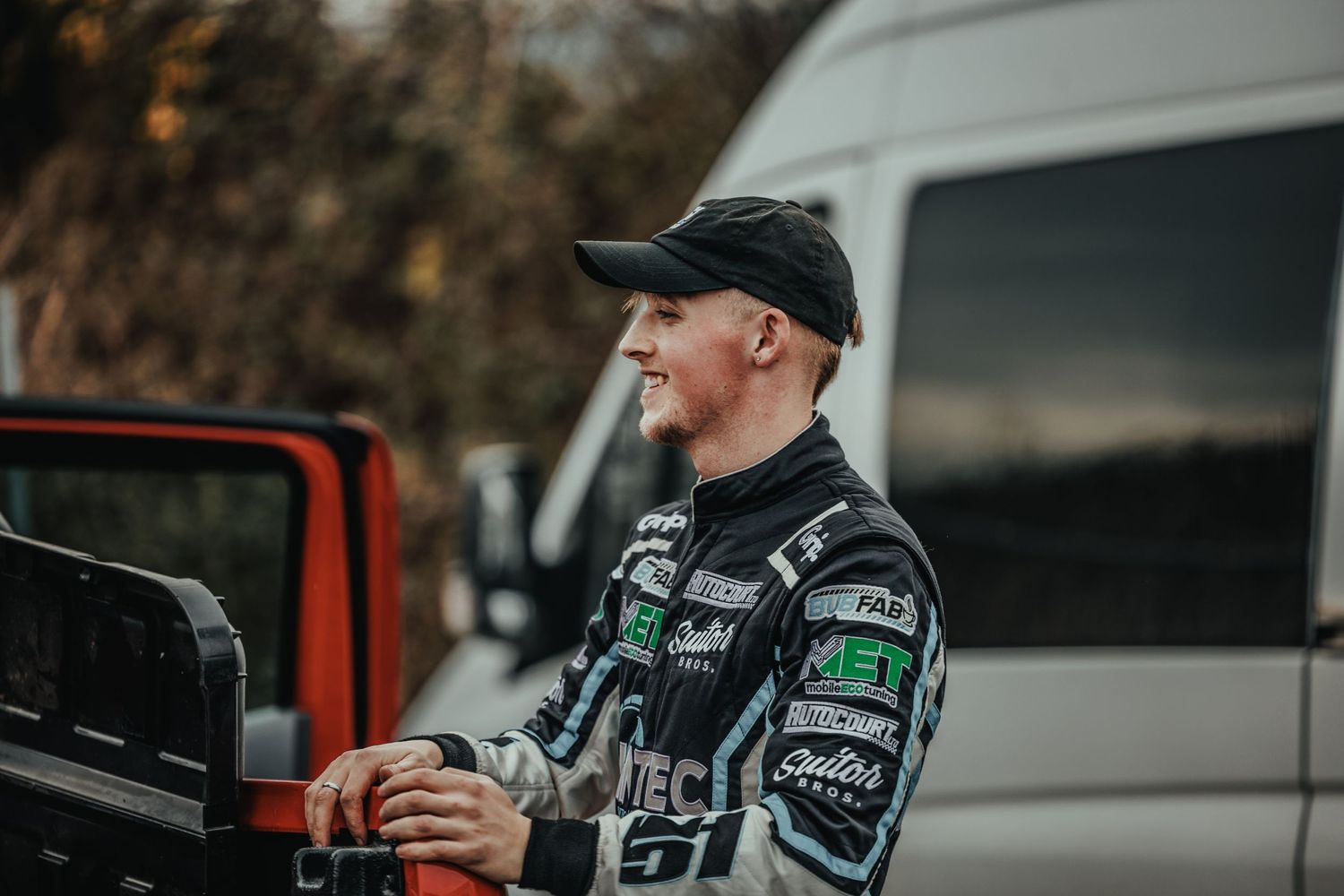 Racing driver in team uniform standing near parked vehicles at competition venue.