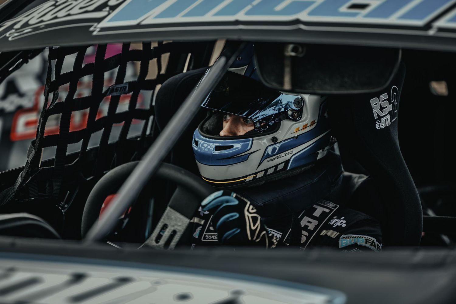 Racing driver in helmet visible through car window netting.