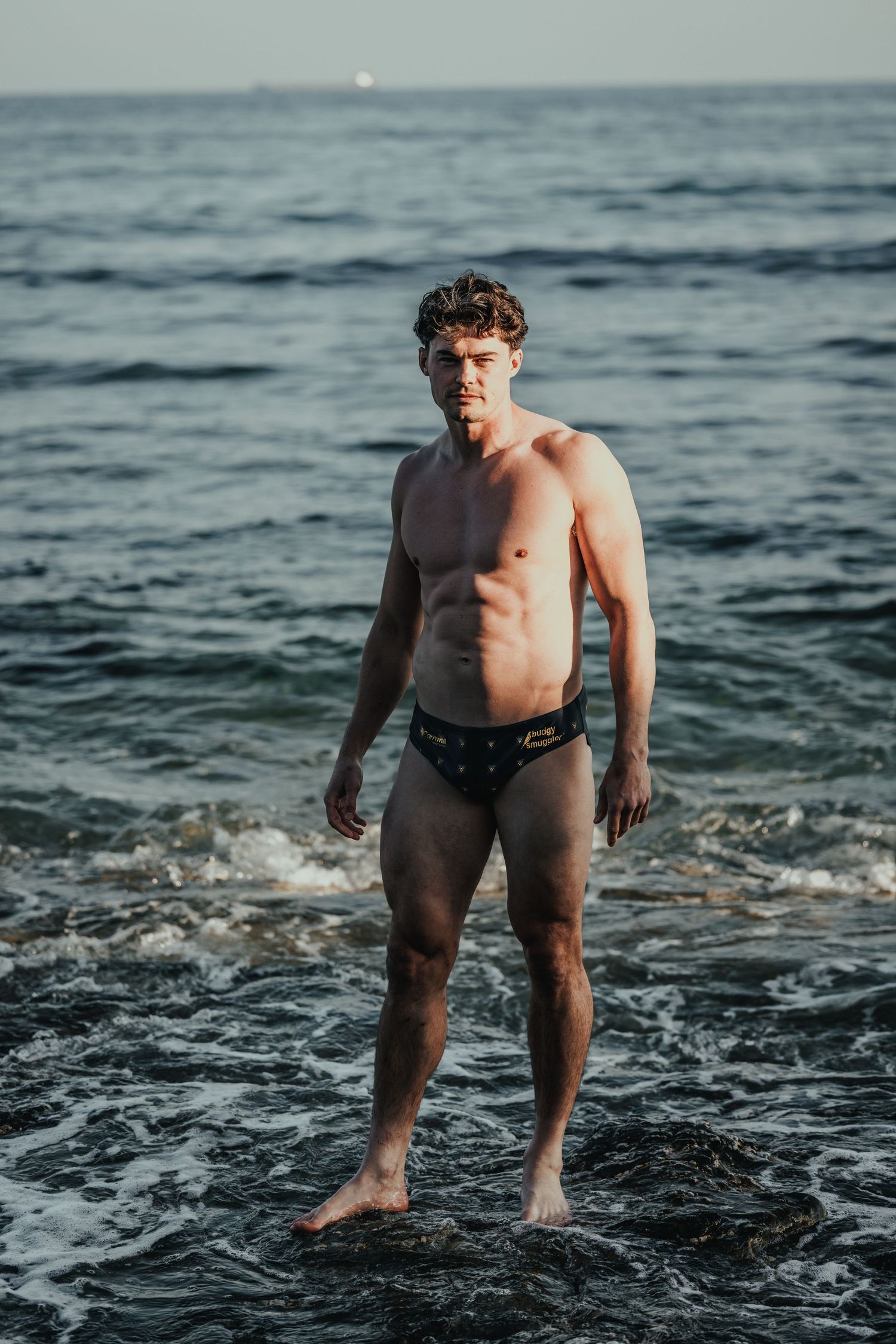 Person in dark swimwear poses confidently in ocean surf at dusk.
