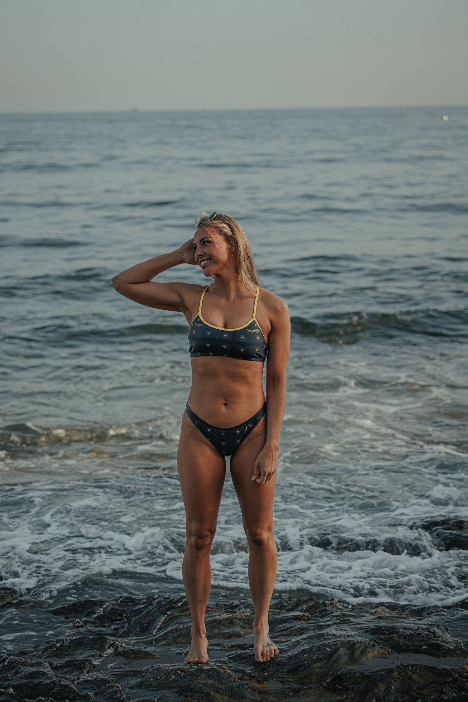 Person in dark bikini poses against dramatic ocean waves during sunset.