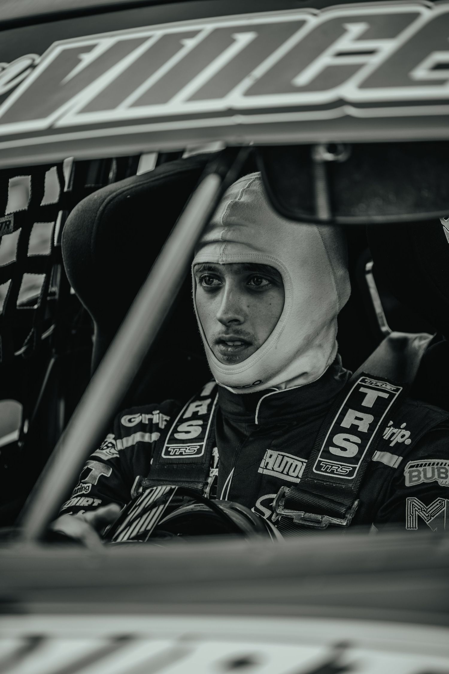 Race car driver in full safety gear and white helmet visible through vehicle window.