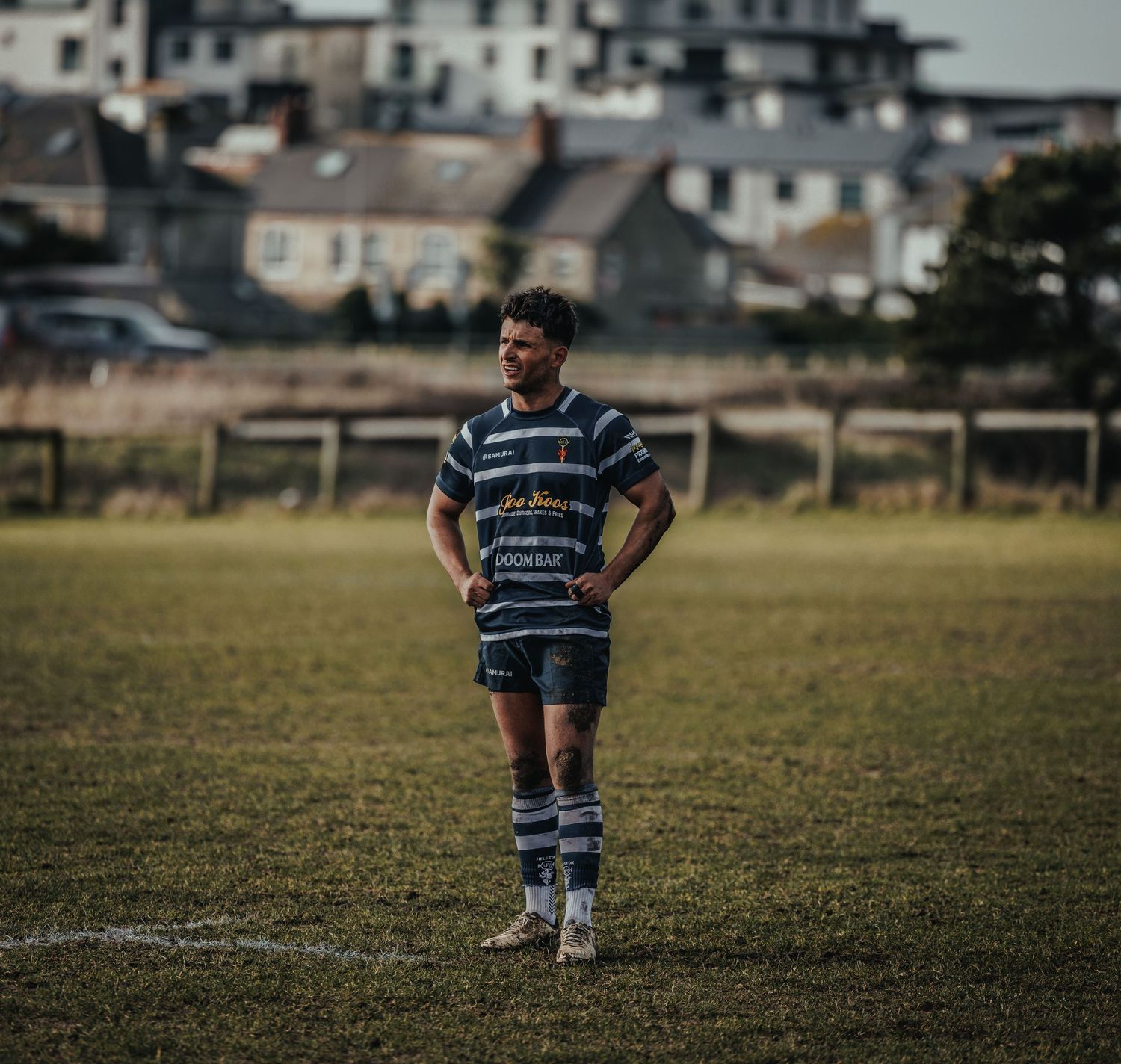 Rugby player in striped jersey stands alone on field with houses in background.