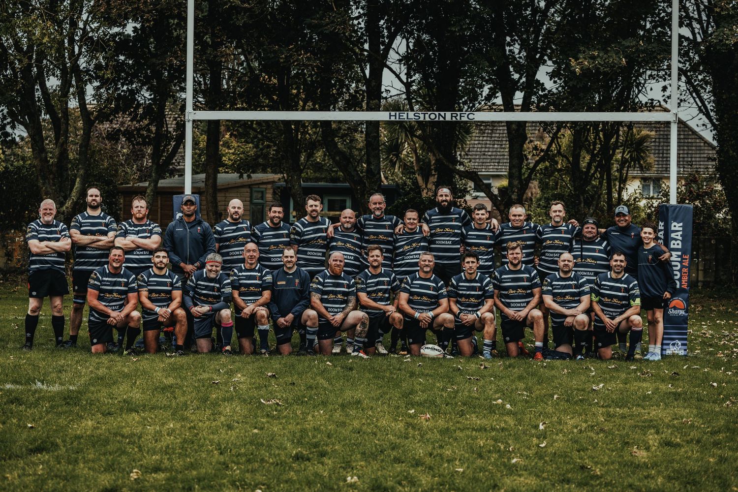 Rugby team poses in striped jerseys on a grass field with goalposts and autumn trees in the background.