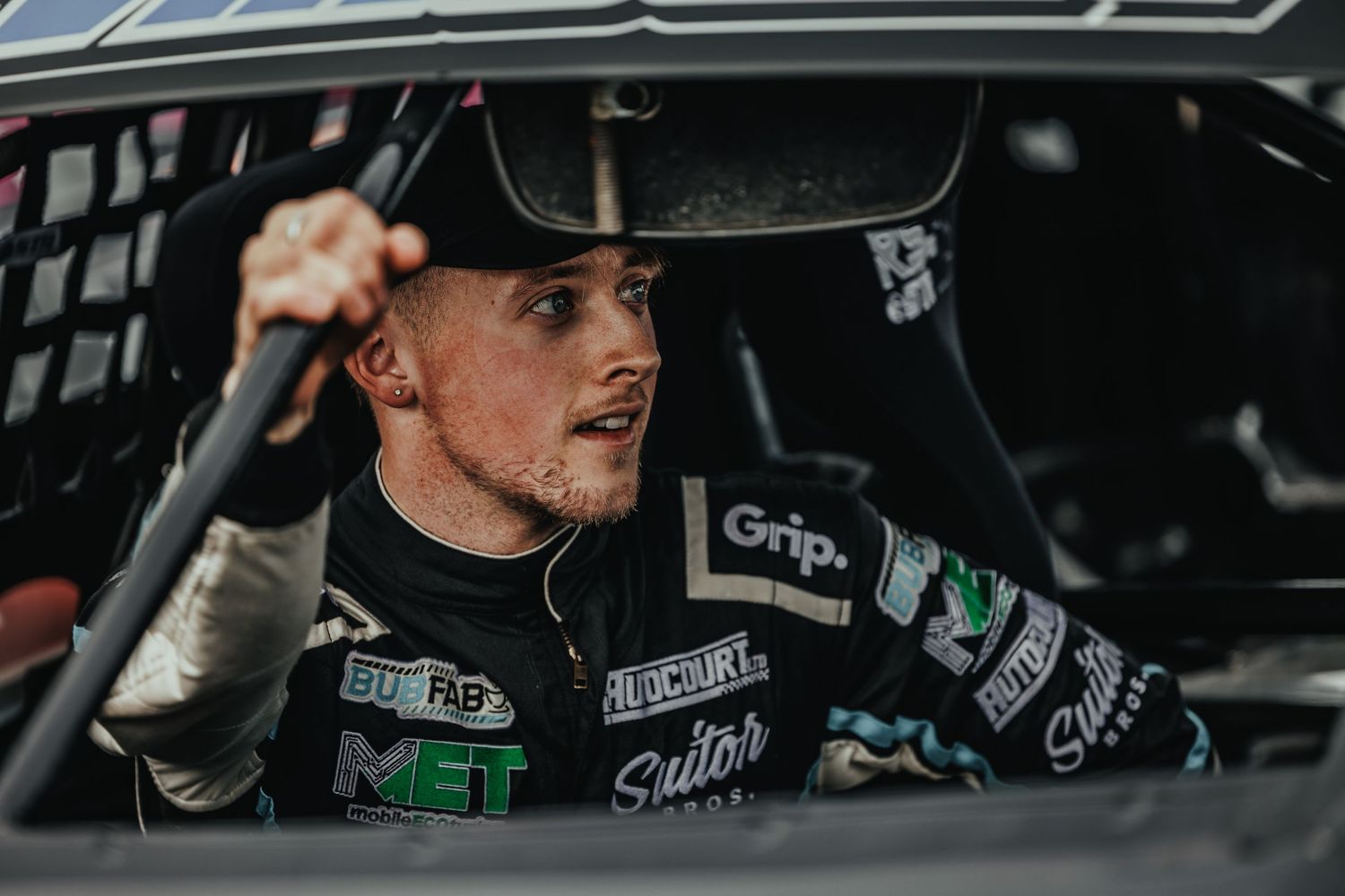 Race driver in sponsored suit and cap inside competition vehicle cockpit.