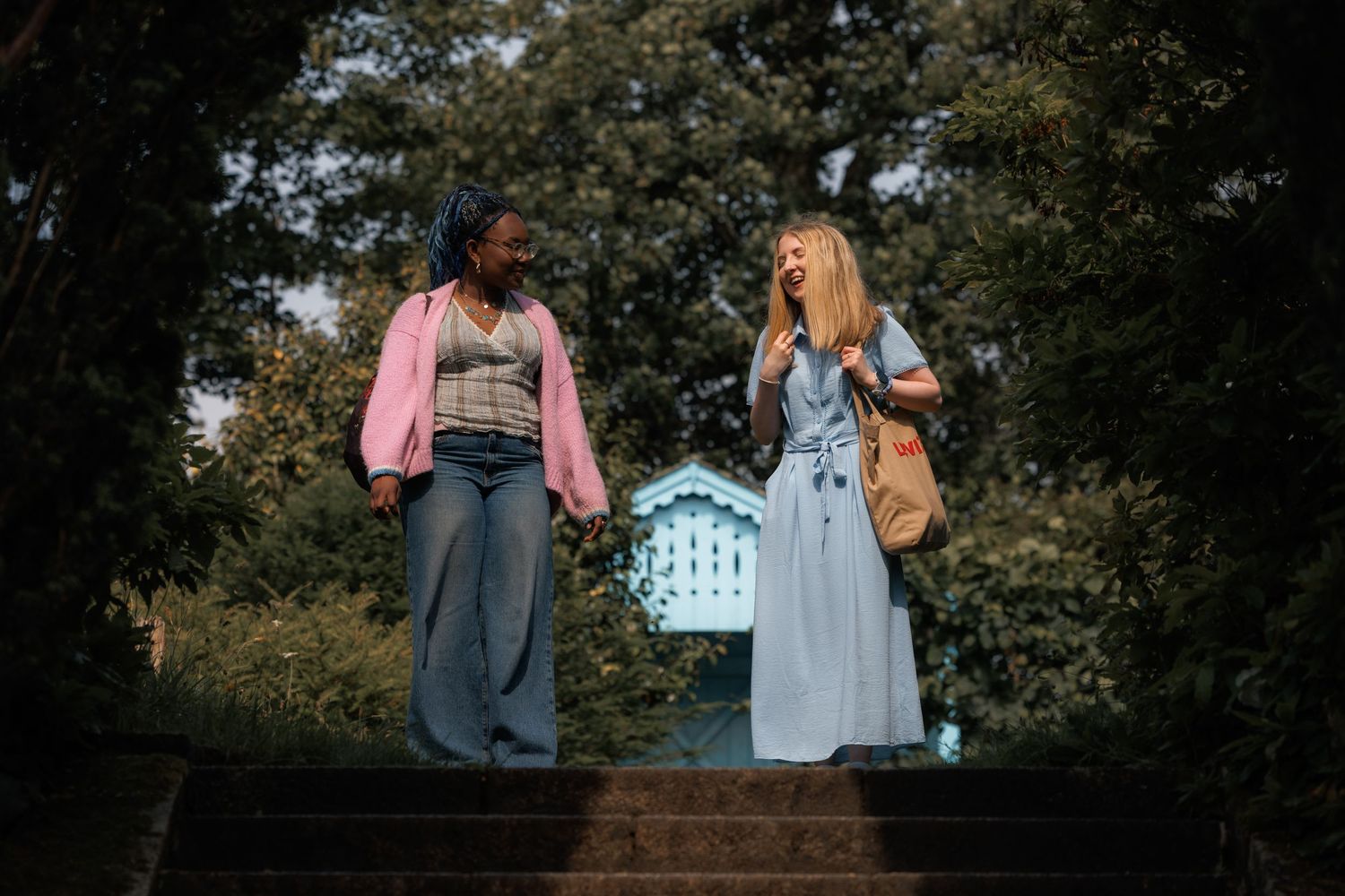 Two women standing on steps in a lush park, one in pink jacket and wide-leg jeans, other in blue dress.