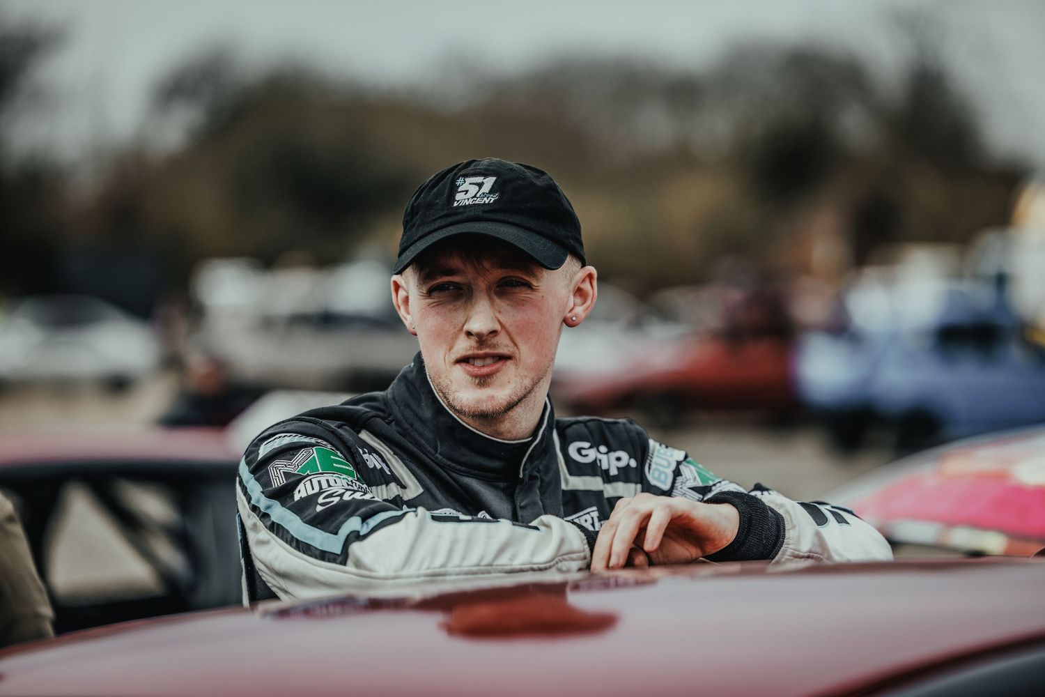 Race car driver in black cap and racing suit leaning against vehicle at motorsports event.