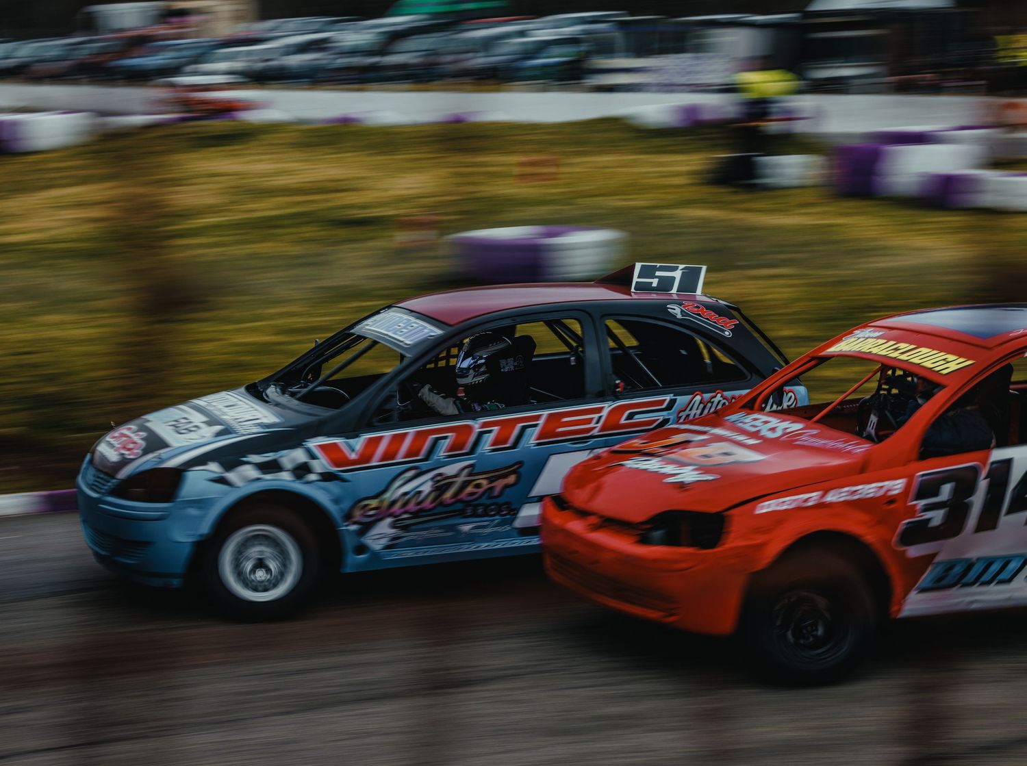 Two stock cars race side by side on a dirt track with motion blur effect.