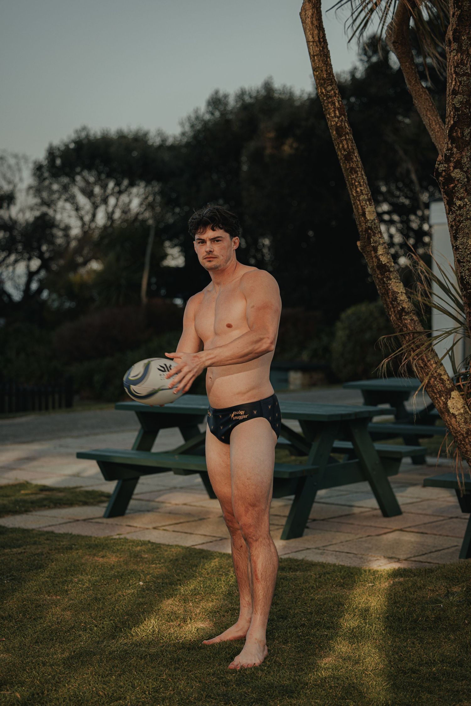 Person in black swimwear holding rugby ball near picnic table at sunset.