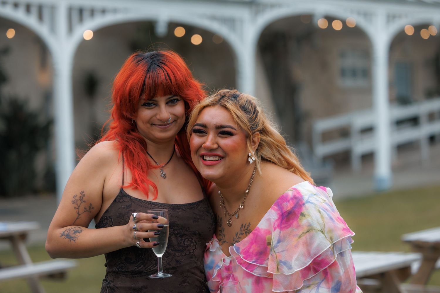 Two women smiling together outdoors at an evening event, one with red hair holding a wine glass.