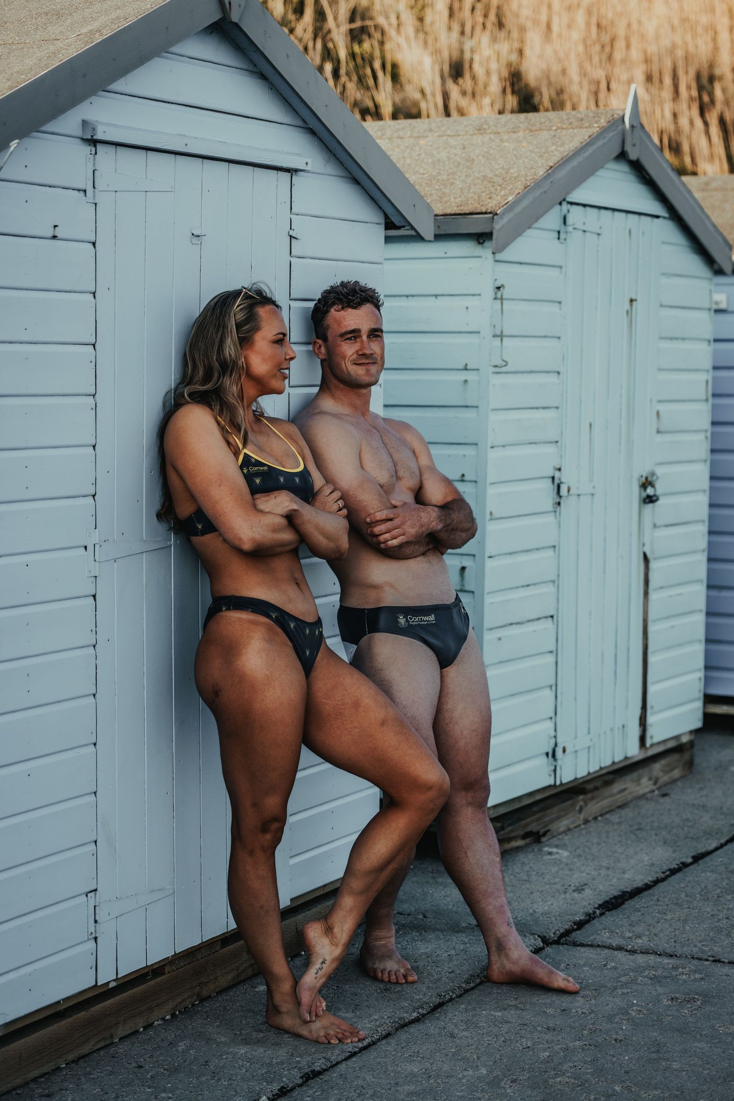 Athletic couple posing in swimwear against pale blue beach huts.