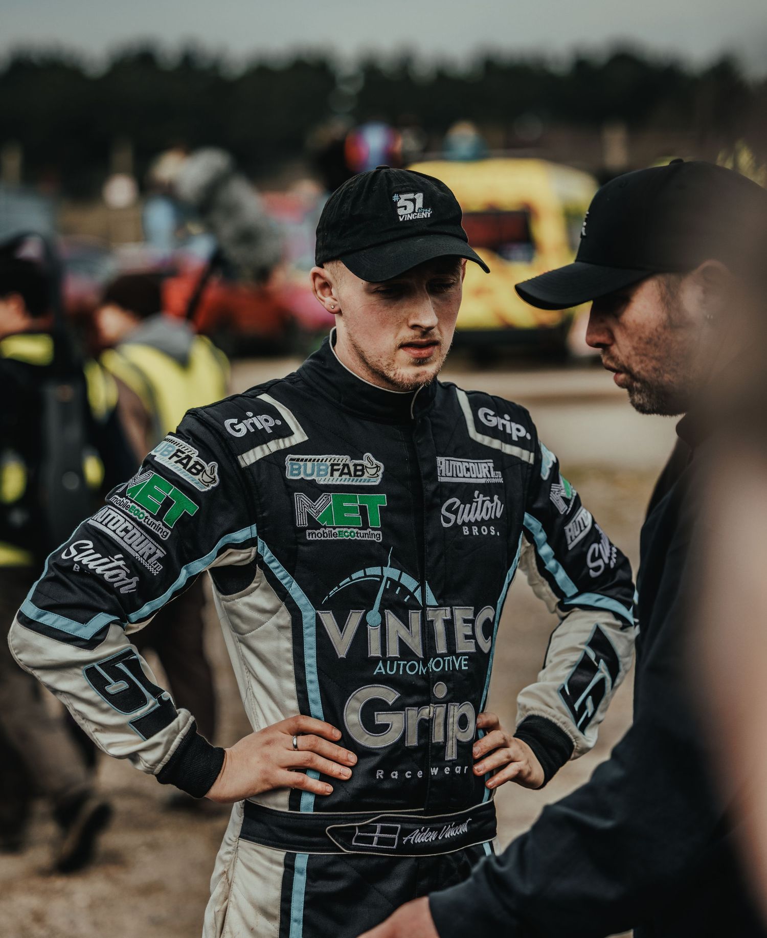 Two motorsport competitors discussing strategy trackside while wearing racing suits.