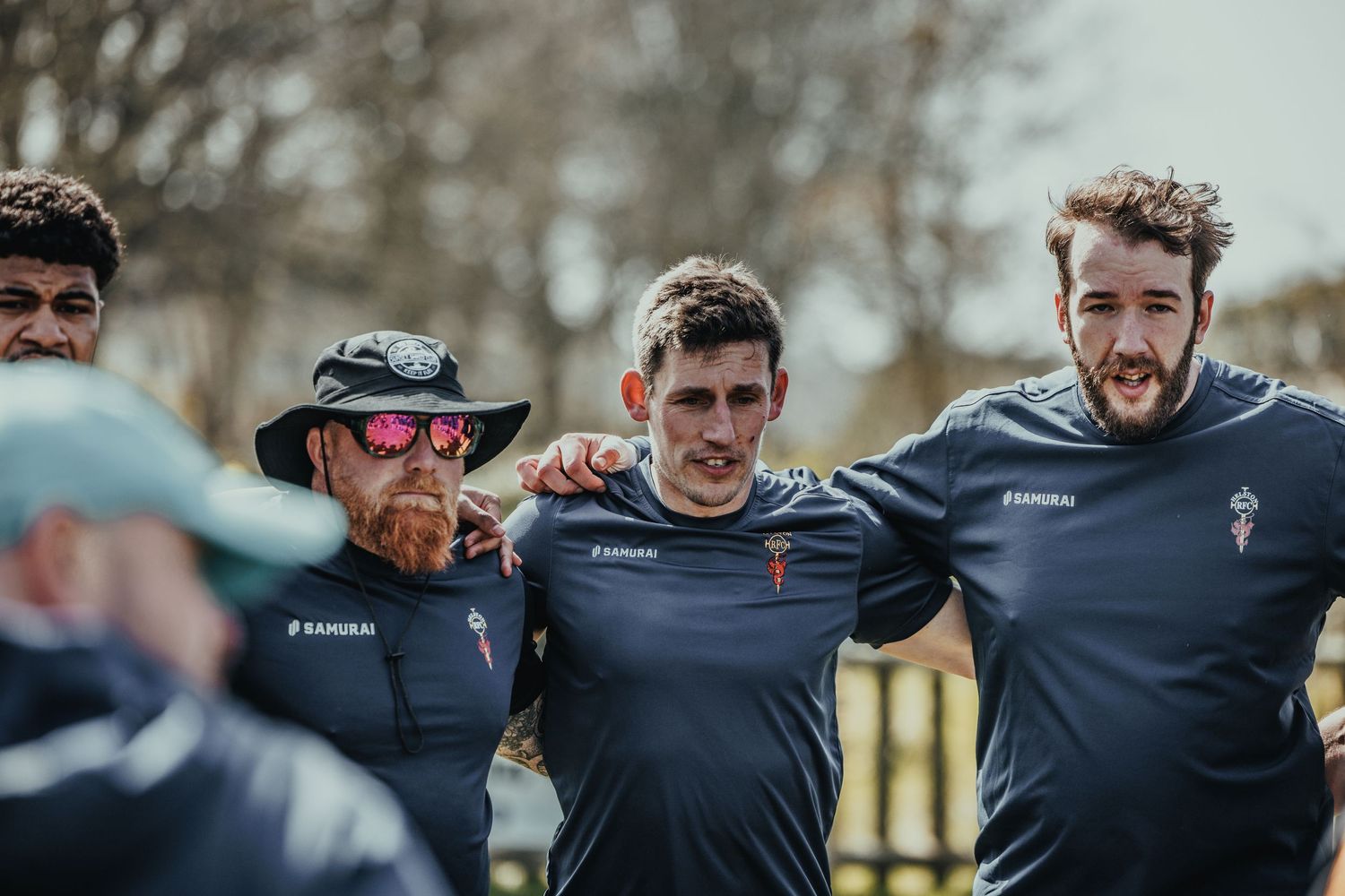Rugby team members huddle together wearing navy training jerseys during practice.