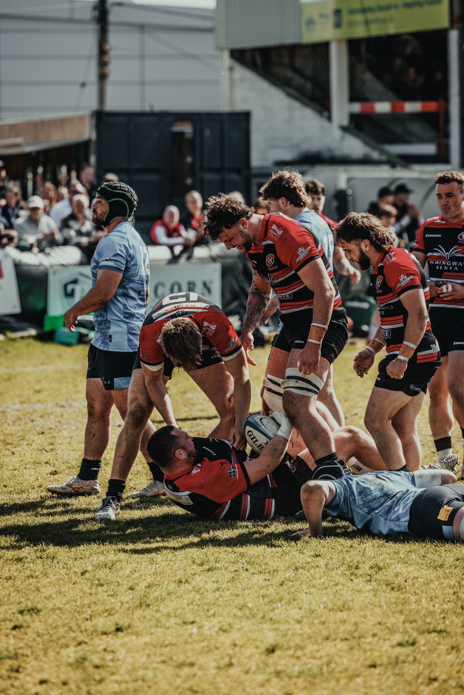 Rugby players in red and black jerseys compete for the ball in an intense match, with spectators watching from the stands.