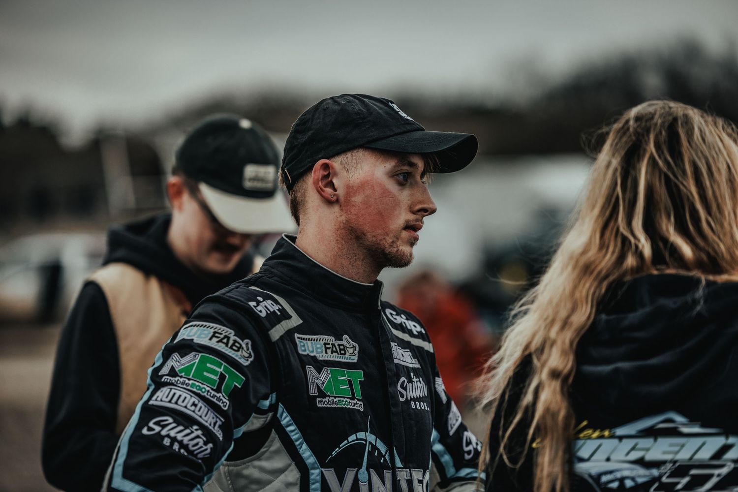 Race car driver in a black sponsored uniform having a conversation at a motorsports event.