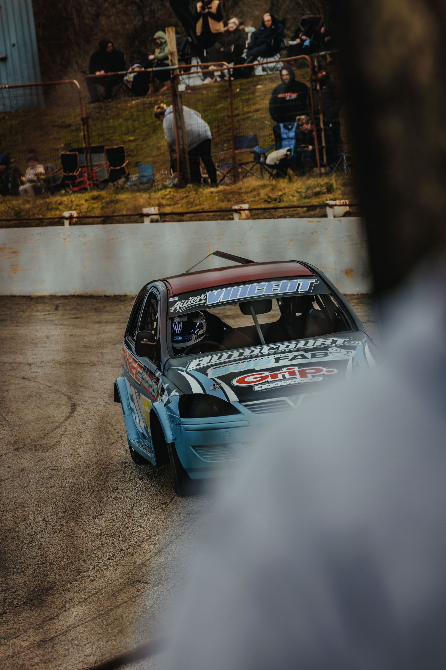 Race car competes on dirt track with spectators watching from elevated viewing area.
