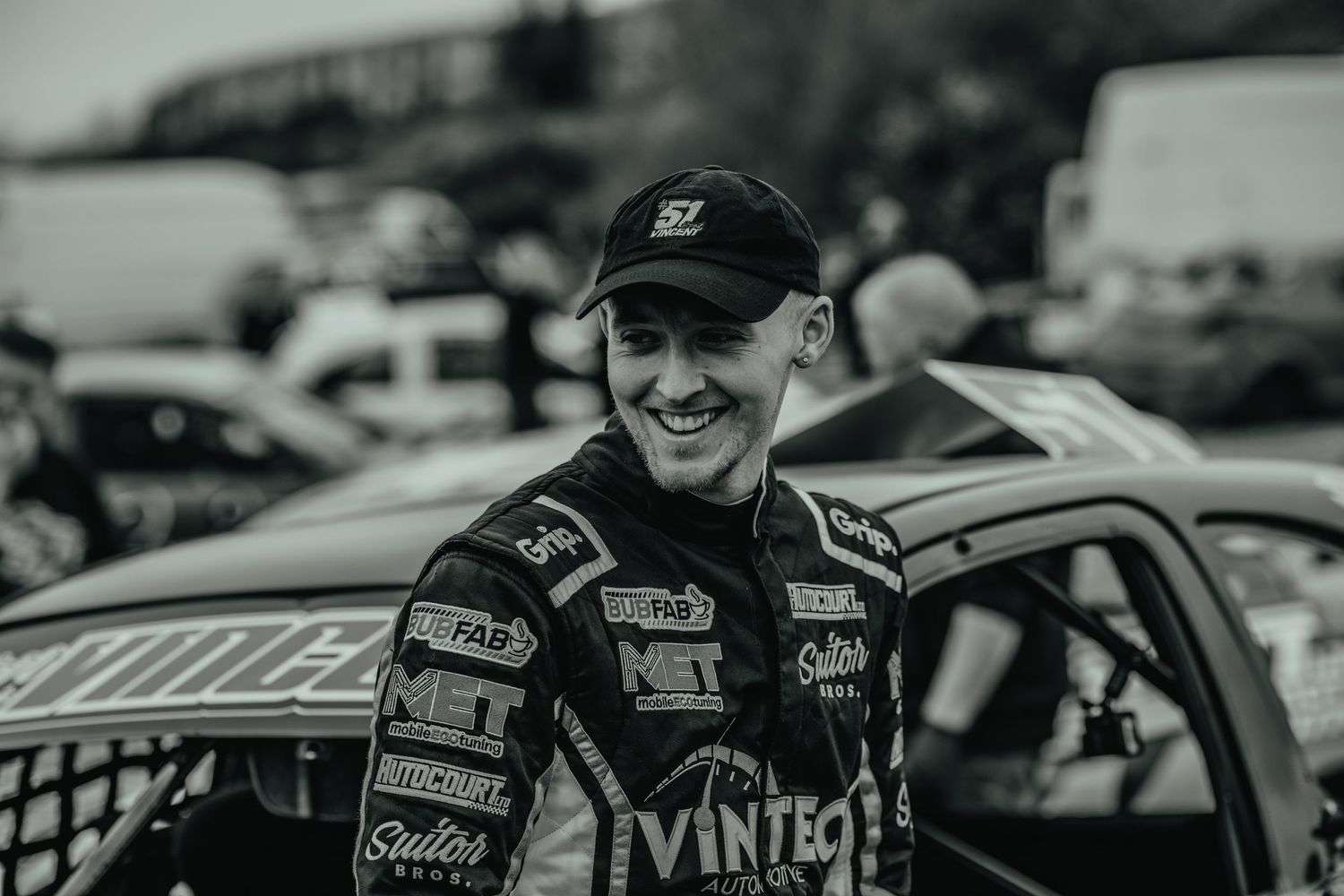 Monochrome portrait of racing driver in Vintec sponsored uniform smiling next to car.