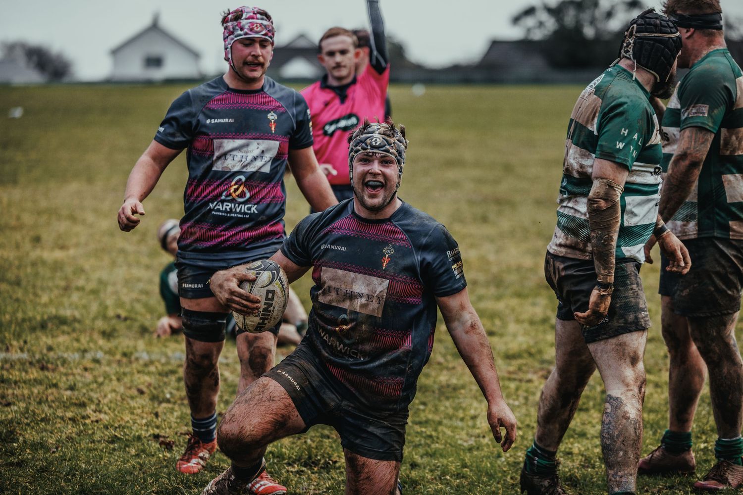 Muddy rugby players celebrate during an intense match on a grass field, with players in dark and green jerseys.