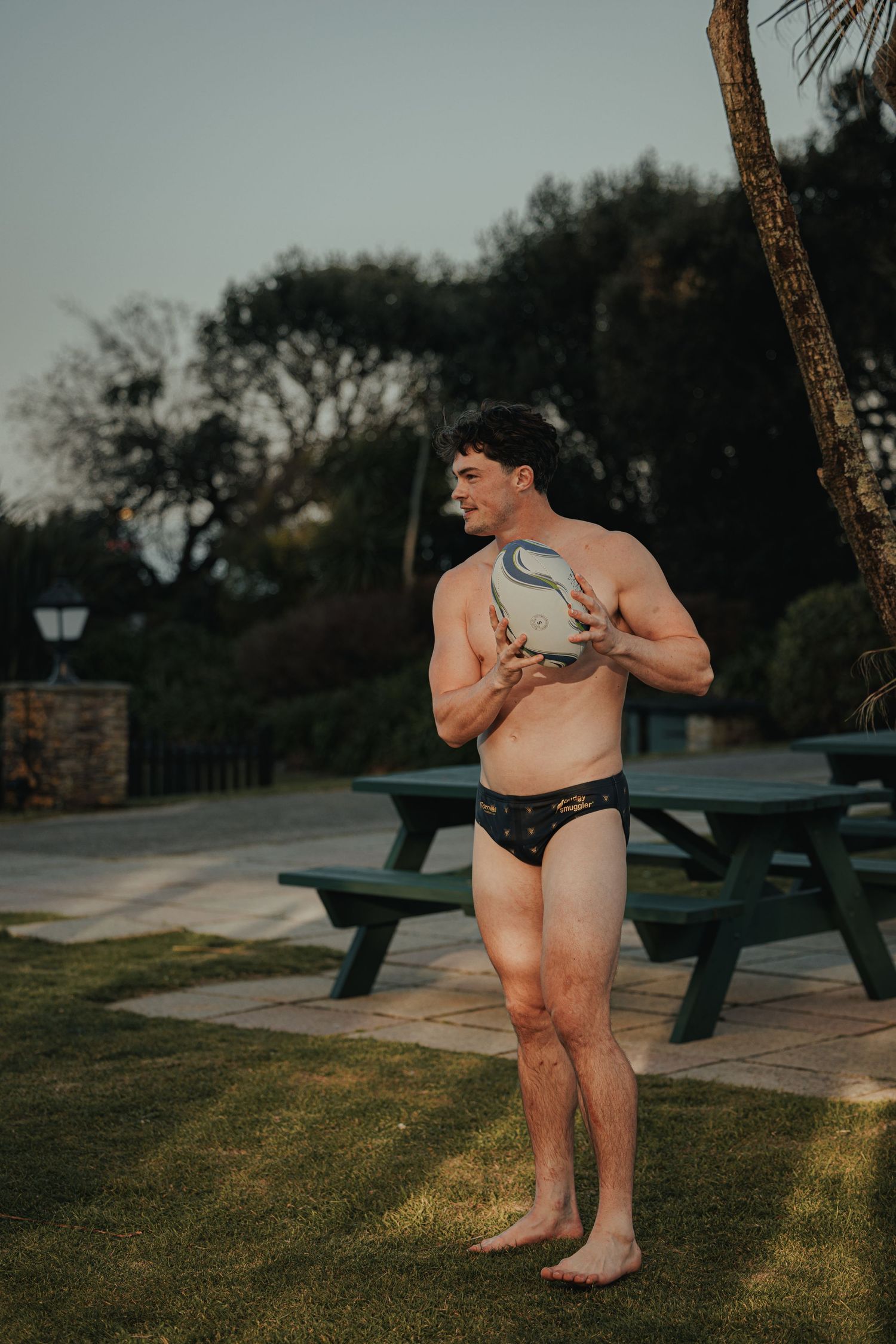 Person in black swimwear holds rugby ball while standing near picnic table.