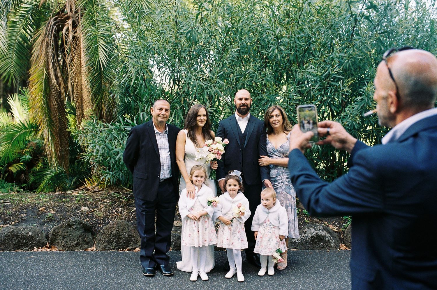 Wedding party posing for photo with bride, groom, flower girls, and guests in front of lush green foliage.