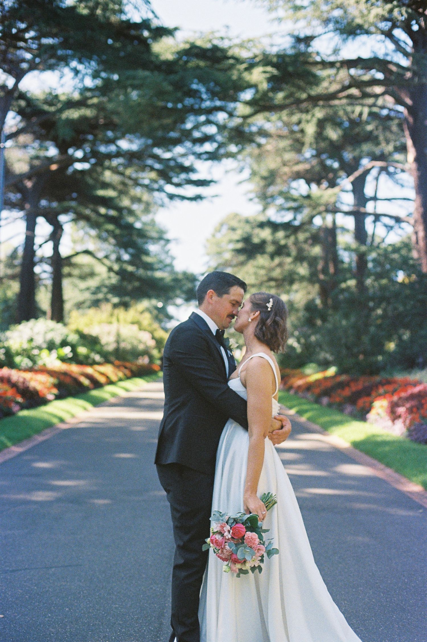 Bride and groom share a romantic moment on a tree-lined path, bride holding a pink bouquet in a white gown.