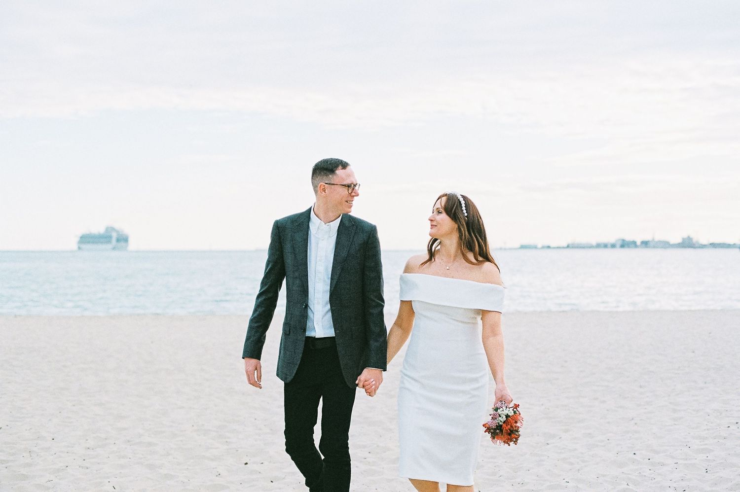 Couple in wedding attire walking hand-in-hand on a sandy beach, bride holding a bouquet, ocean in the background.