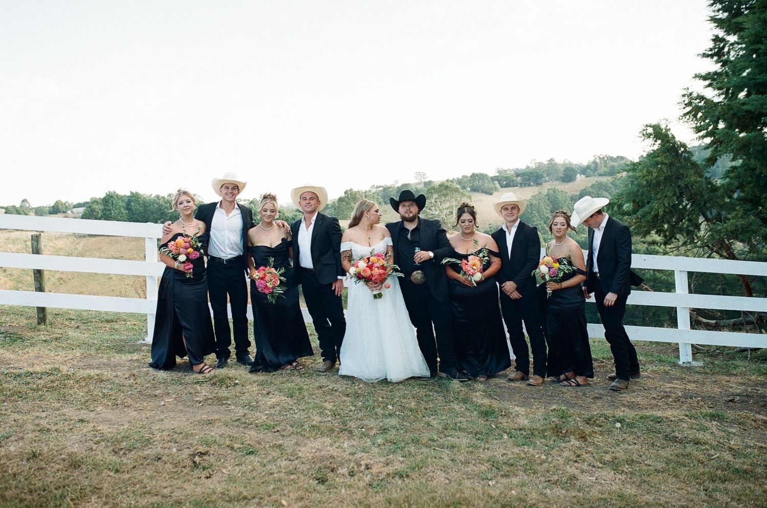 Panoramic sequence of wedding party members sitting and posing on a grassy hillside by white fences during sunset.