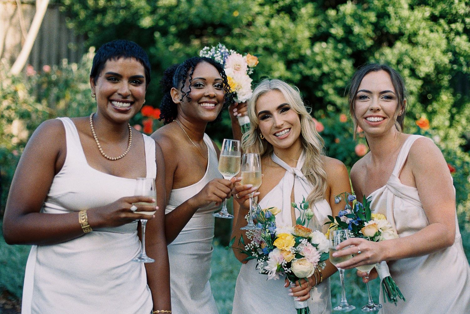 Four bridesmaids in white dresses toasting with champagne glasses in a lush garden wedding celebration.
