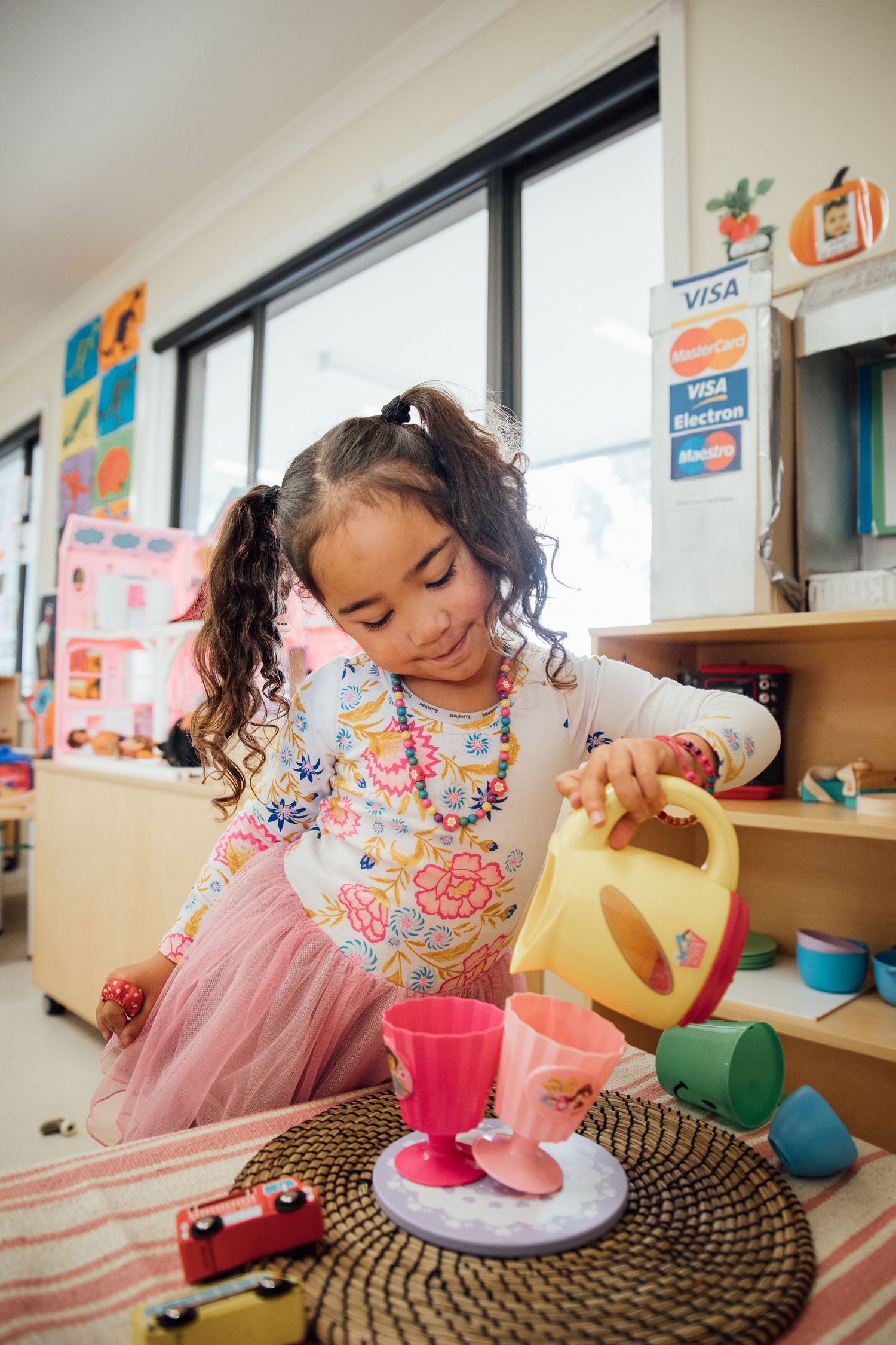 A young child in a floral dress pours tea from a yellow teapot into colorful cups during playtime.