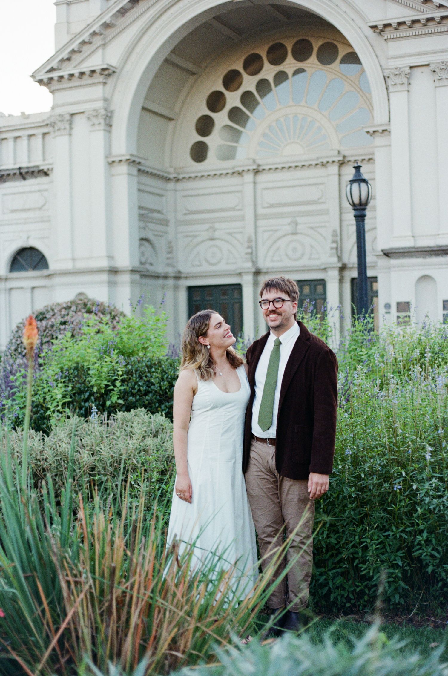 A couple in wedding attire stands smiling in a lush garden before a grand white neoclassical building.
