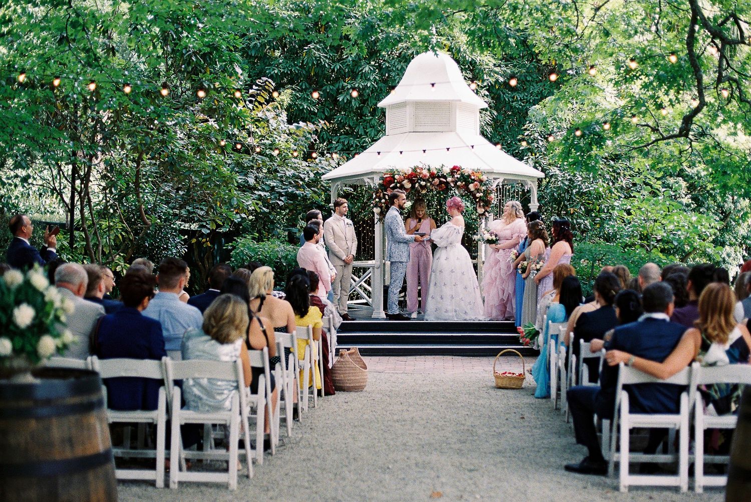 Outdoor wedding ceremony at a white gazebo surrounded by lush trees, with guests seated in white chairs on a gravel path.