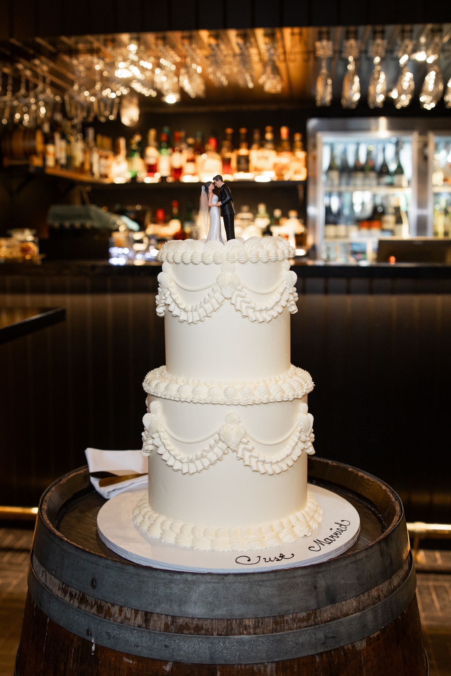 Three-tier white wedding cake with bride and groom topper, displayed on a wooden barrel in an elegant bar setting.