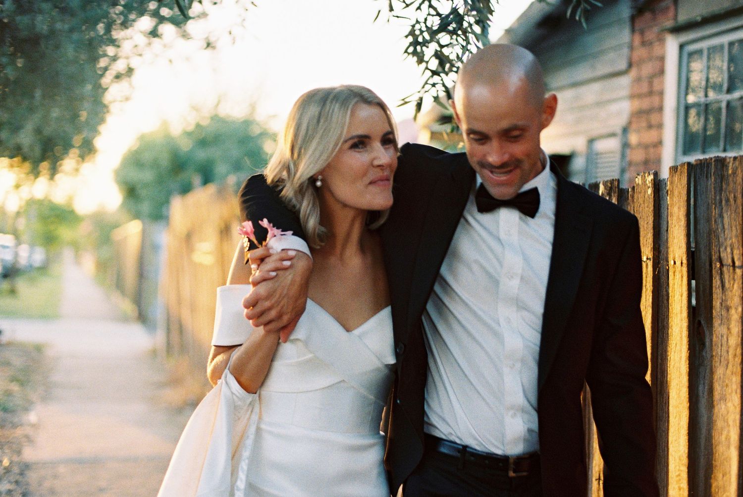 A bride in a white gown and groom in a black tuxedo share a romantic moment outdoors near a wooden fence.