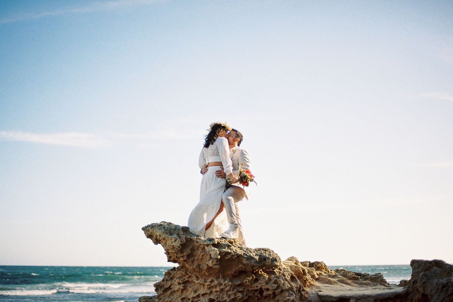 Bride and groom embracing on rocky seaside cliff with ocean waves and blue sky backdrop.