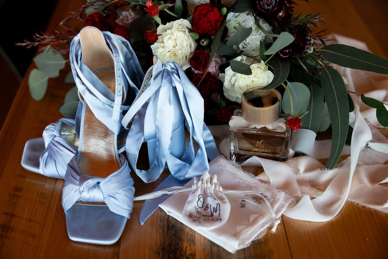 Light blue wedding shoes and accessories displayed with red and white floral decorations on wooden surface.