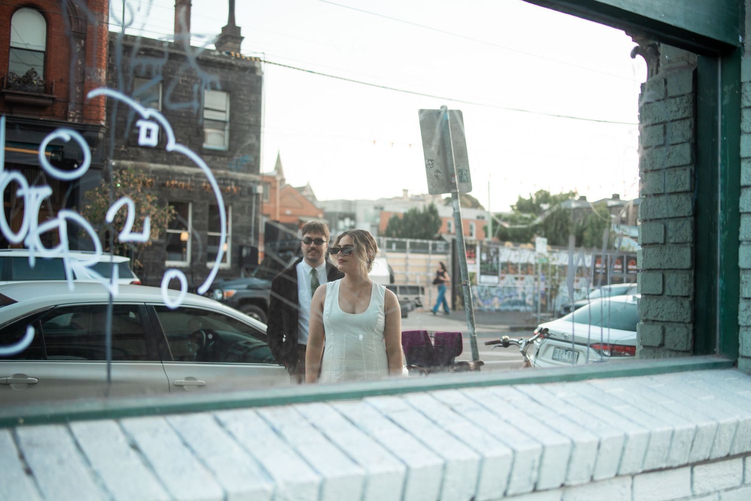 Woman in white tank top walking past a graffiti-covered storefront window on a urban city street.