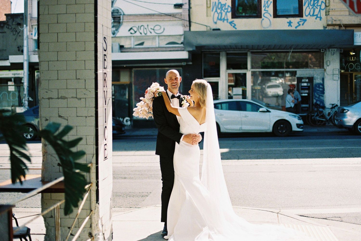 Bride and groom embracing on a city street corner, bride in white gown with veil, groom in black tuxedo.