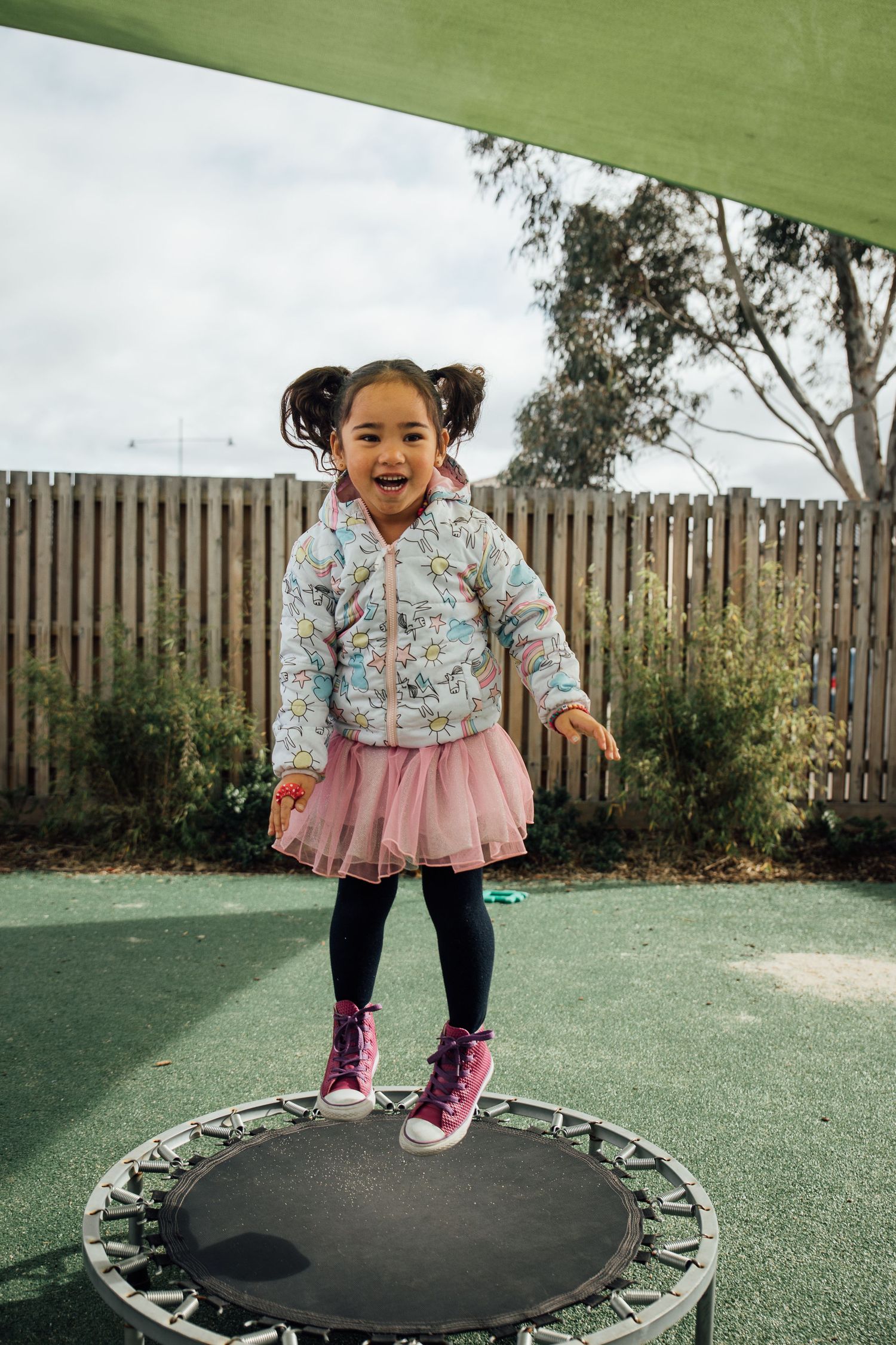 A child wearing a floral jacket and pink tutu skirt jumps on a small trampoline in a backyard play area.