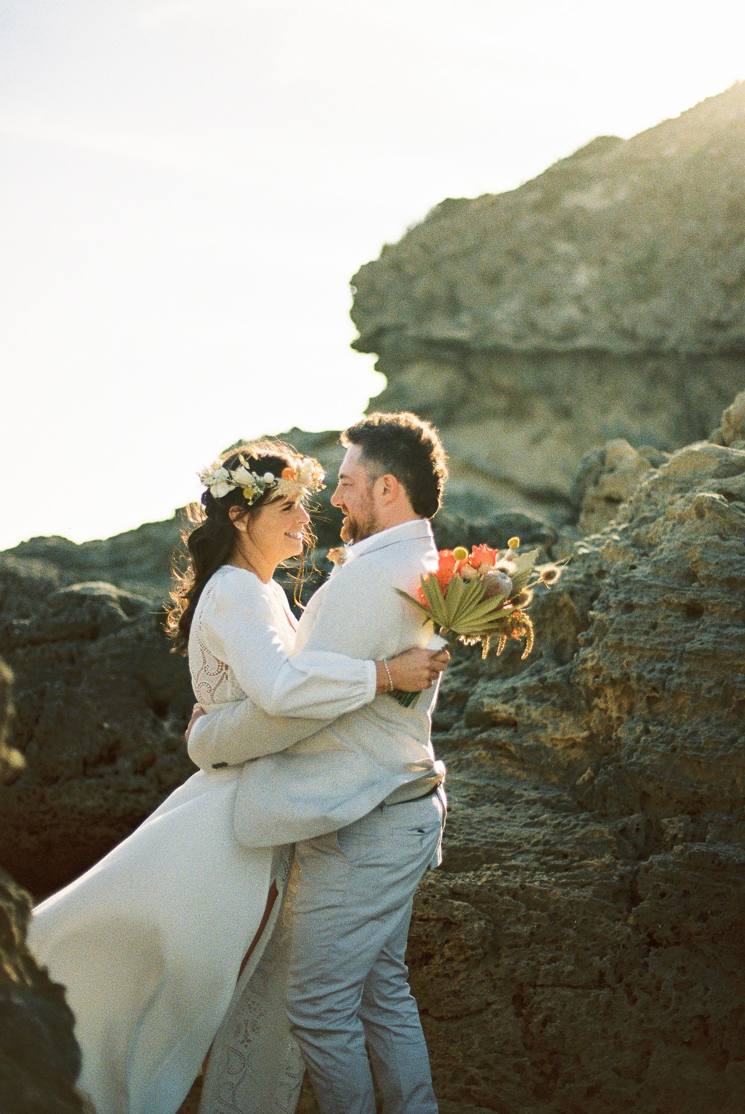 Couple in wedding attire embracing near rocky coast, bride wearing floral crown holding bouquet, backlit by golden sunlight.