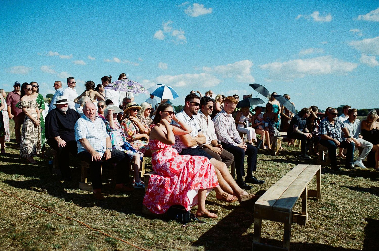 Outdoor wedding ceremony guests seated on benches under blue sky, woman in pink floral dress prominent in crowd.