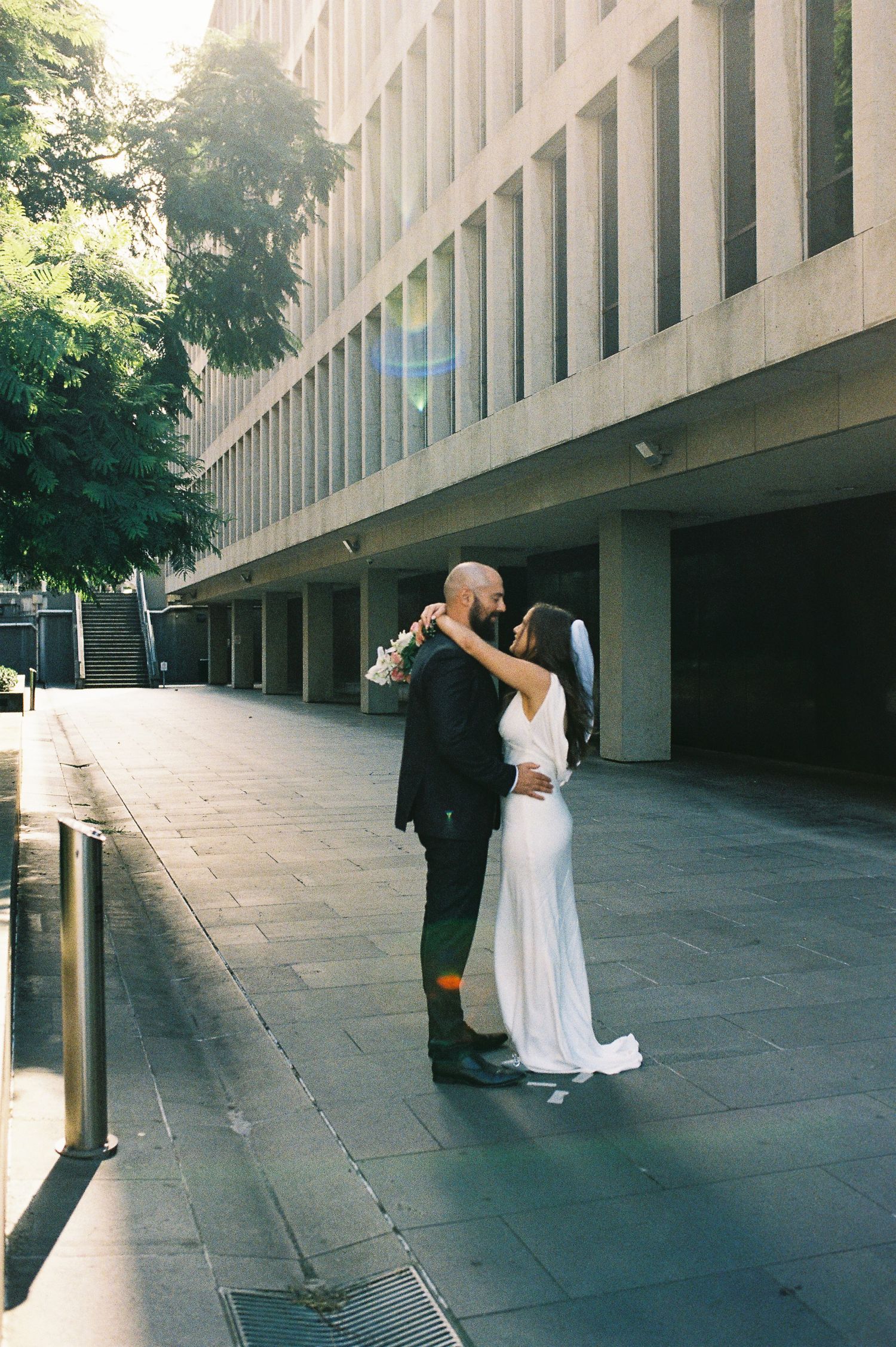Bride and groom embracing on urban sidewalk outside modern building, bride in white gown, groom in dark suit.