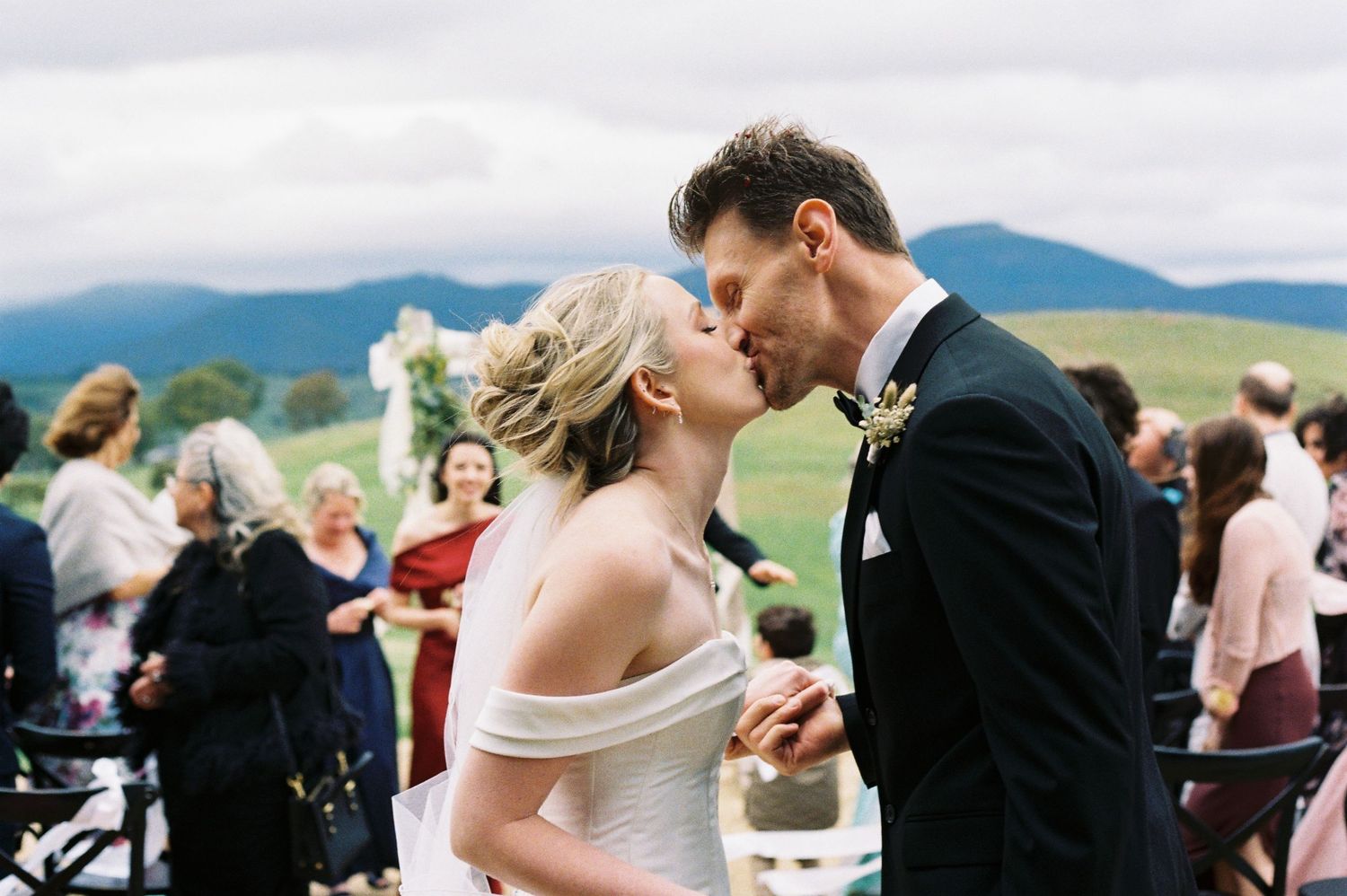Bride and groom share first kiss at outdoor wedding ceremony with mountain views and guests celebrating behind them.